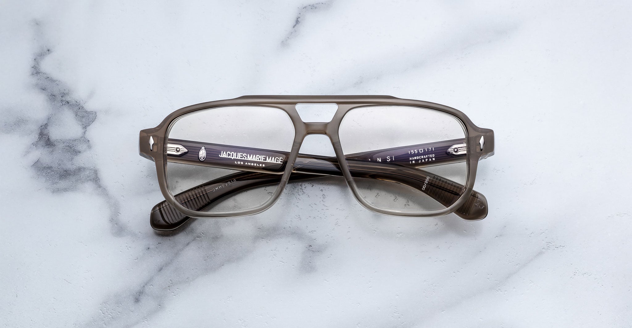 The Jacques Marie Mage Evans eyeglasses by Jacques Marie Mage feature an oversized gray acetate frame with thick rims and rectangular lenses, shown resting on a white marble surface.
