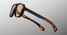 Close-up of Jacques Marie Mage Evans sunglasses: oversized acetate frame, black fronts, brown lenses, and tortoiseshell-patterned temples by Jacques Marie Mage, showcased on a gray gradient background.