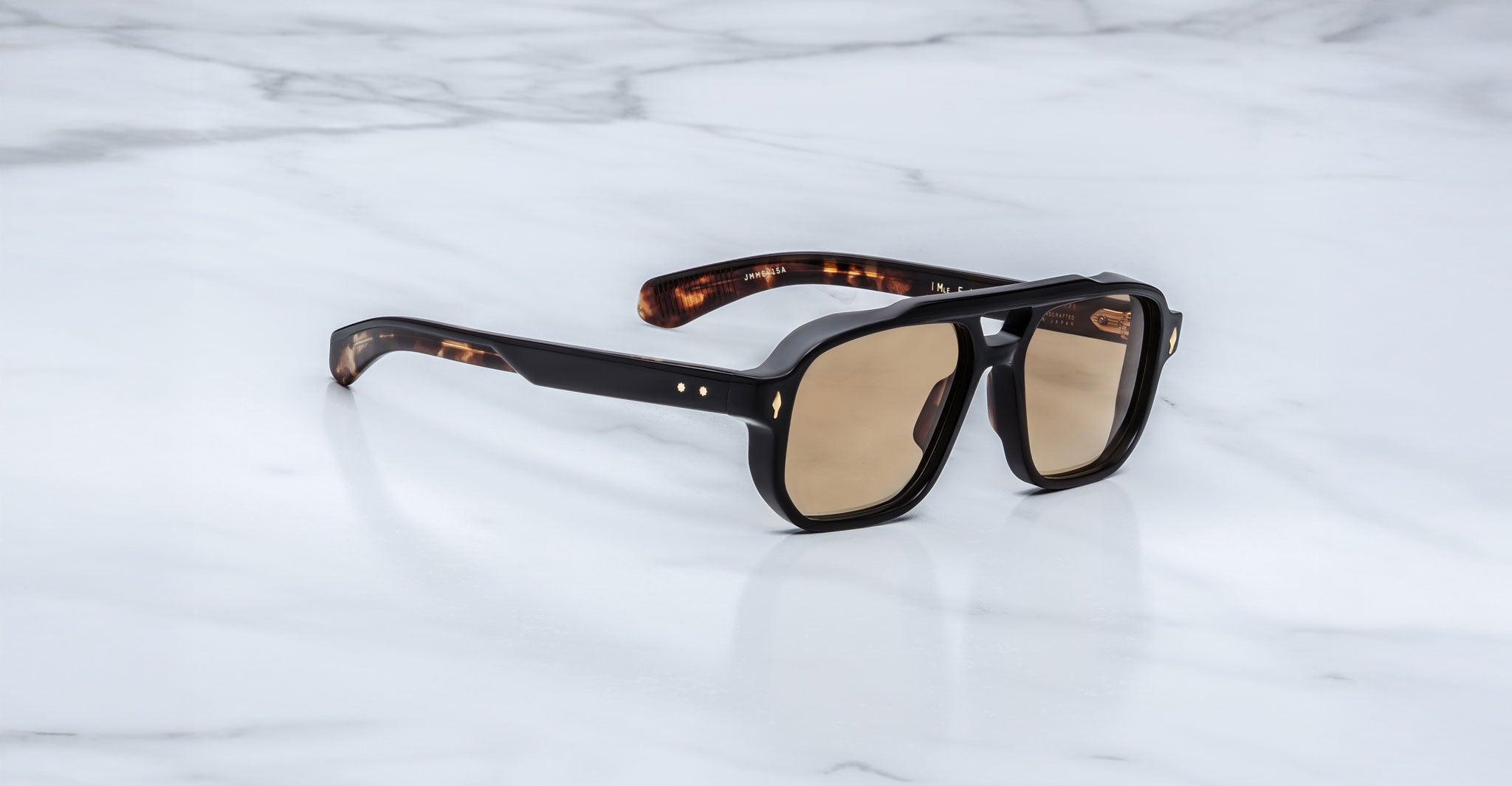 The Jacques Marie Mage Evans sunglasses in Noir 15 feature brown-tinted lenses and thick, dark tortoiseshell oversized acetate frames, resting on a white marble surface.