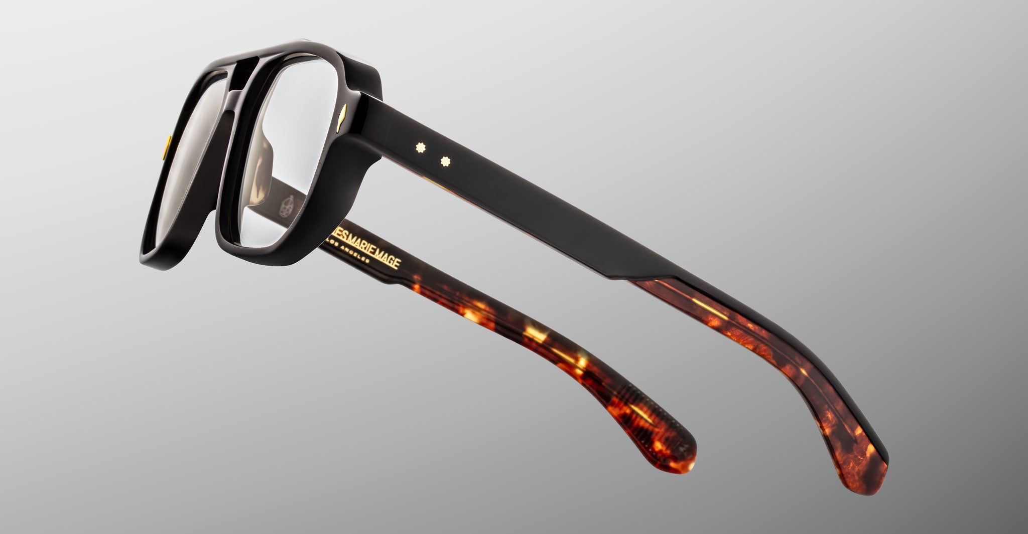 The Jacques Marie Mage Evans features handcrafted optical glasses with thick black oversized acetate frames and tortoiseshell-patterned temple arms, set against a gray gradient background.