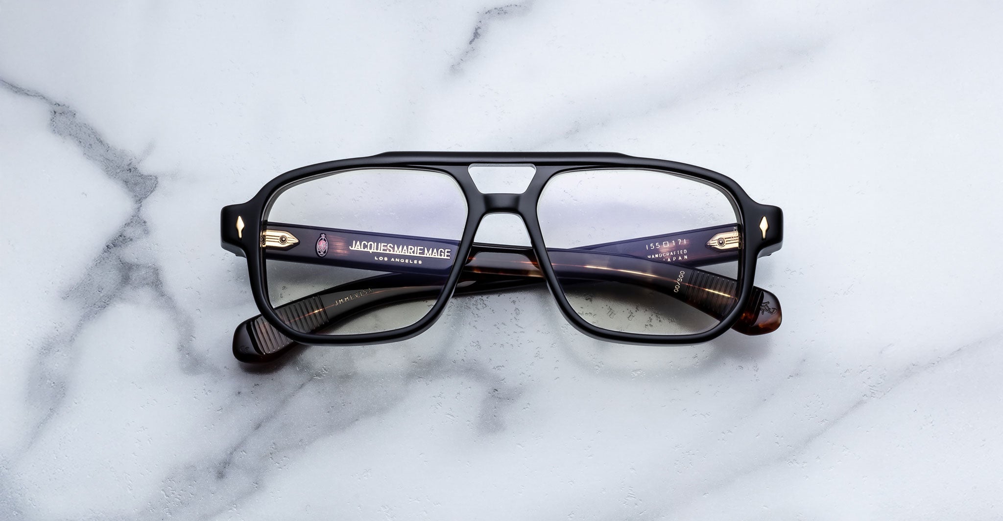 A pair of Jacques Marie Mage Evans black rectangular eyeglasses with an oversized acetate frame rests on a white marble surface.