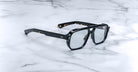 The Jacques Marie Mage Evans eyeglasses feature thick, oversized black acetate frames with clear lenses, echoing classic handcrafted designs, and are displayed on a white marble surface.