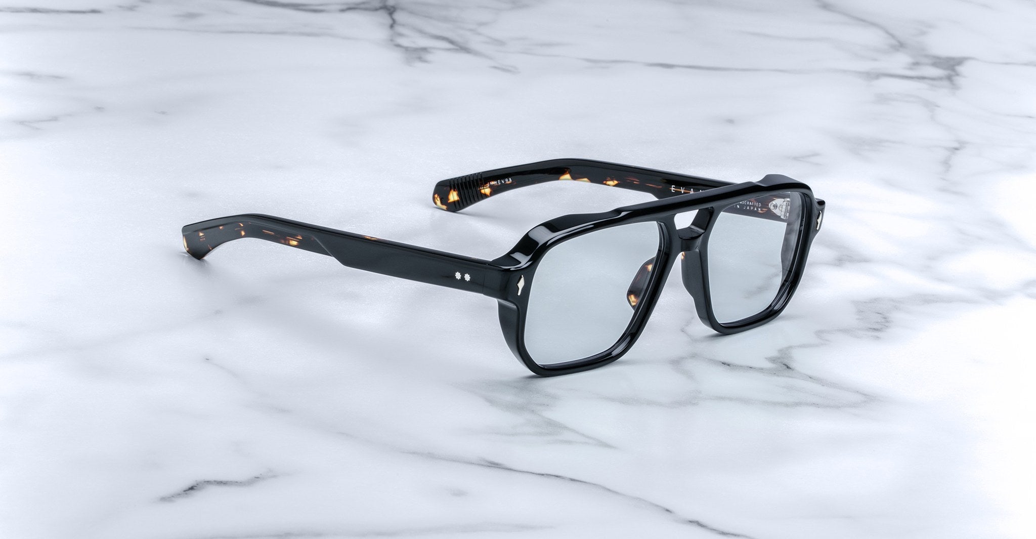 The Jacques Marie Mage Evans eyeglasses feature thick, oversized black acetate frames with clear lenses, echoing classic handcrafted designs, and are displayed on a white marble surface.