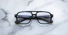 Jacques Marie Mage Evans by Jacques Marie Mage—black square eyeglasses with an oversized acetate frame and clear lenses, arms folded on a white marble surface—a modern handcrafted optical design.