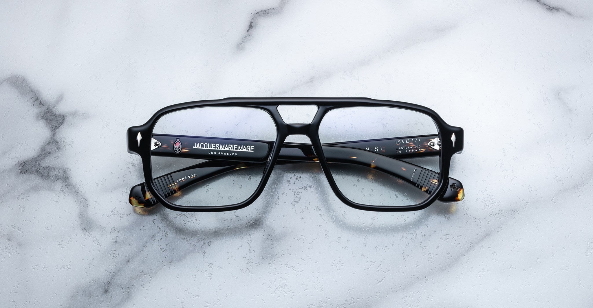 Jacques Marie Mage Evans by Jacques Marie Mage—black square eyeglasses with an oversized acetate frame and clear lenses, arms folded on a white marble surface—a modern handcrafted optical design.