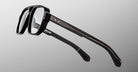 The Jacques Marie Mage Evans are black, thick-rimmed handcrafted optical glasses with clear lenses and metal-accented temples by Jacques Marie Mage, shown against a gray background.