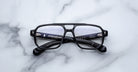 The Jacques Marie Mage Evans by Jacques Marie Mage are black, rectangular, handcrafted optical glasses with clear lenses and an oversized acetate frame, displayed on a white marble surface.