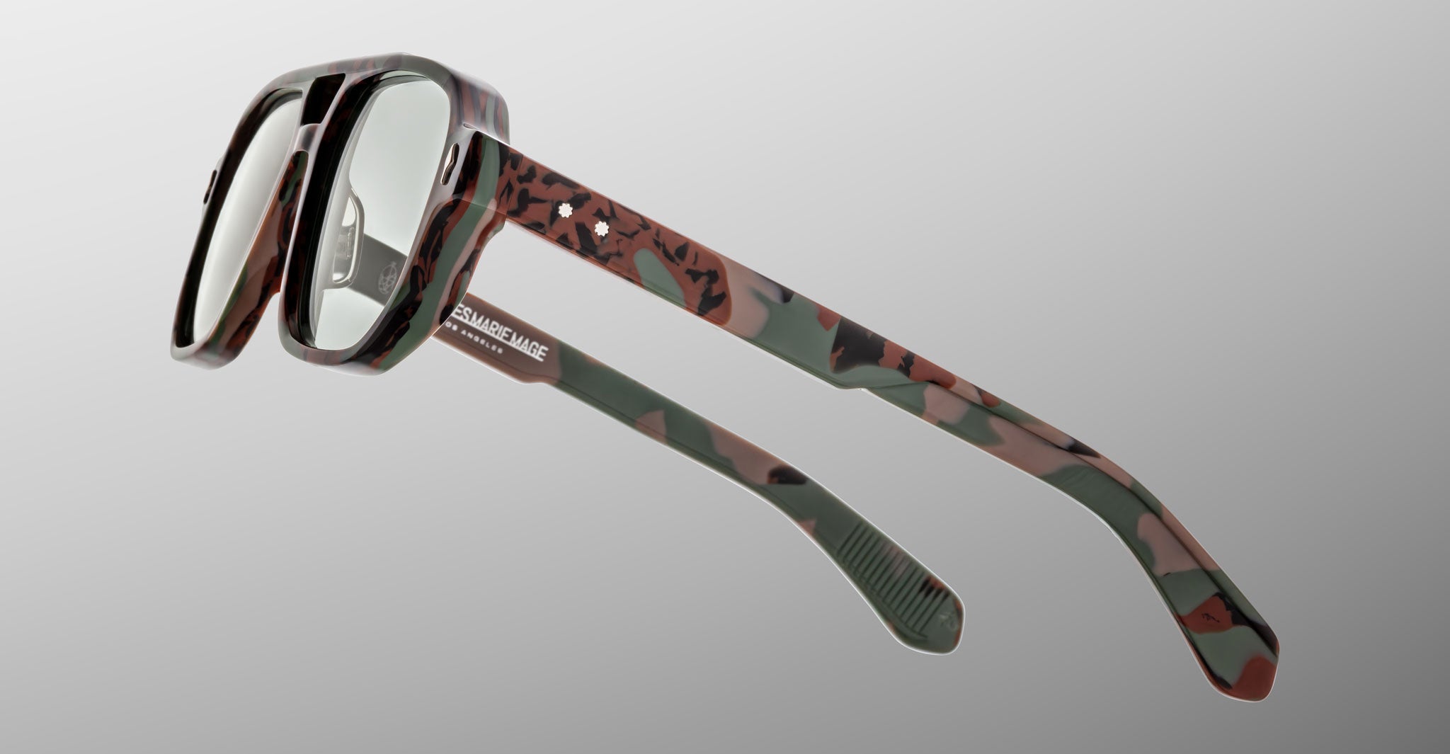 Close-up of Jacques Marie Mage Evans glasses, showcasing handcrafted oversized acetate frames in a camouflage pattern with clear lenses, set against a plain gray background.