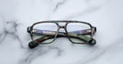 The Jacques Marie Mage Evans optical glasses by Jacques Marie Mage showcase an oversized green camouflage acetate frame with clear lenses, elegantly displayed on a white marble surface.