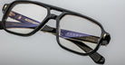 Close-up of Jacques Marie Mage Evans black-rimmed eyeglasses with oversized acetate frames, featuring visible branding on the arms, set against a light grey background.
