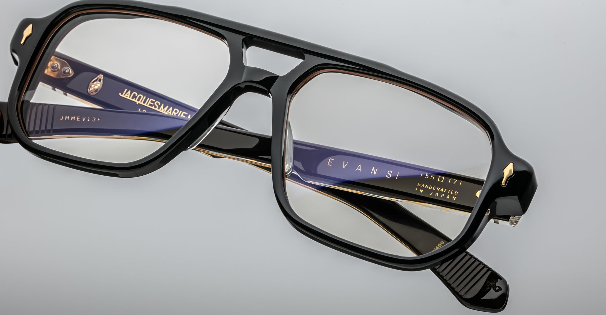 Close-up of Jacques Marie Mage Evans black-rimmed eyeglasses with oversized acetate frames, featuring visible branding on the arms, set against a light grey background.