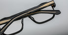 Close-up of Jacques Marie Mage Evans optical glasses, handcrafted with black and gold accents and an oversized acetate frame, shown folded against a plain gray background.