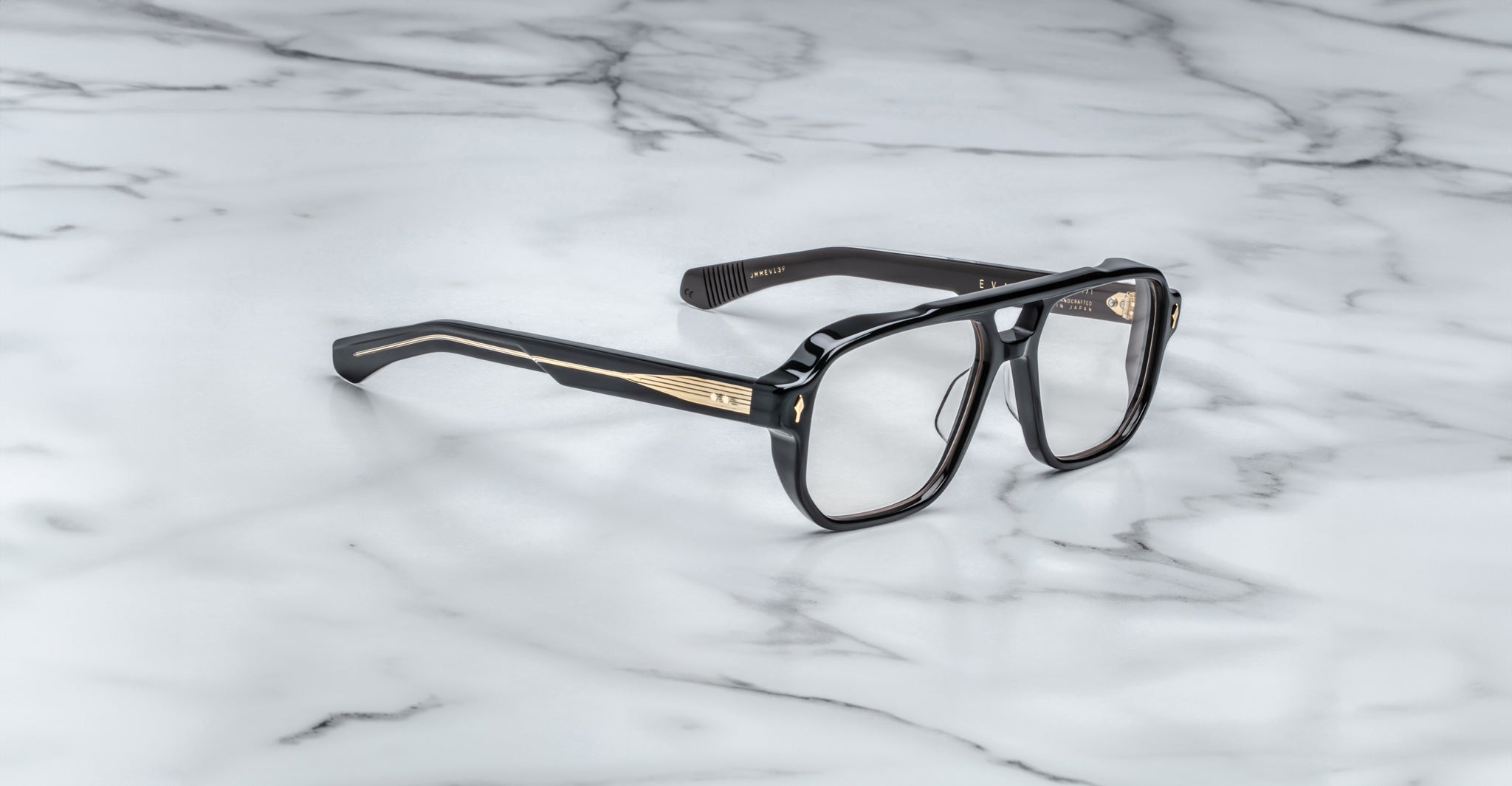 The Jacques Marie Mage Evans handcrafted optical glasses feature an oversized black acetate frame with gold temple detailing, displayed on a white marble surface.