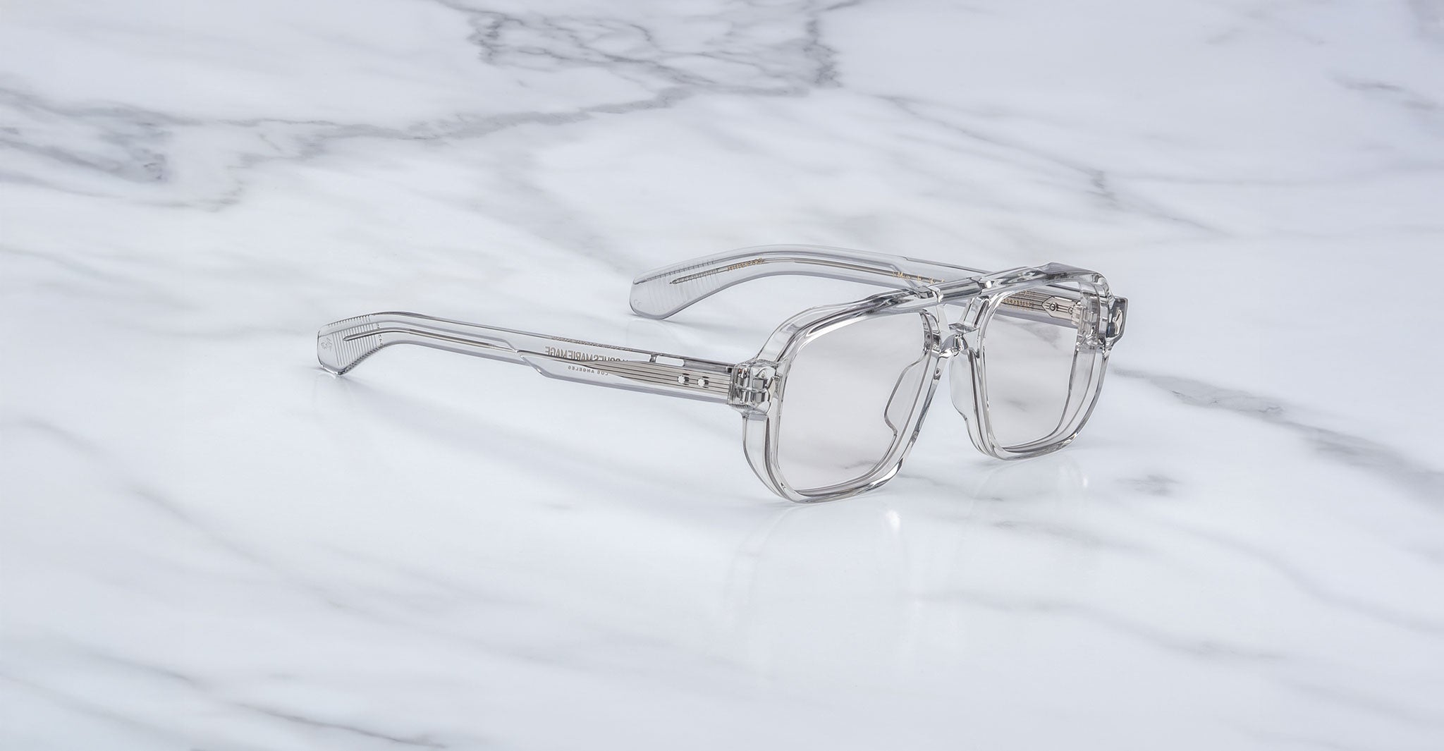 Jacques Marie Mage Evans clear-framed handcrafted optical glasses with transparent lenses are displayed on a white marble surface.