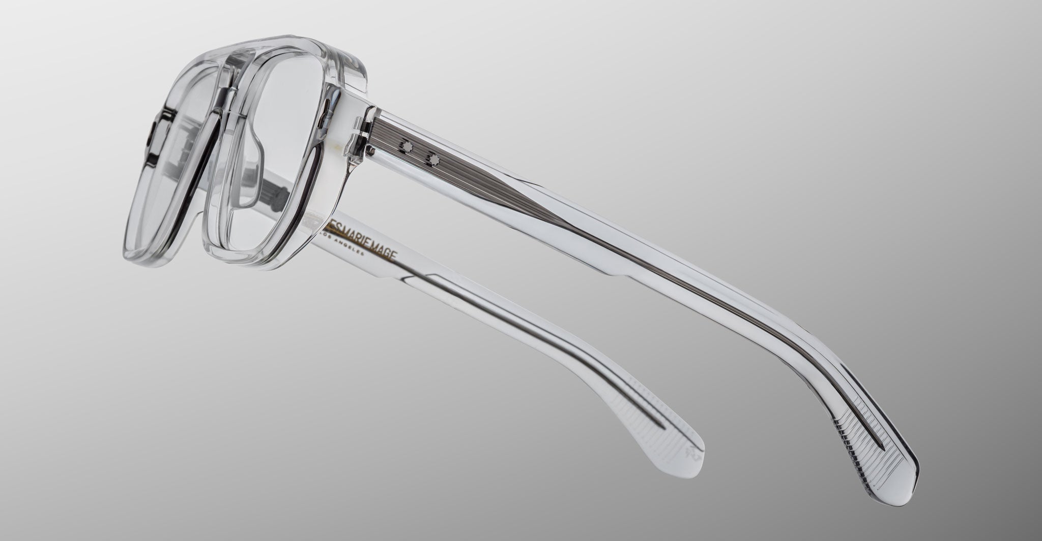 The Jacques Marie Mage Evans optical glasses feature oversized clear acetate frames with translucent arms and visible metal hinges, shown against a neutral gradient background.