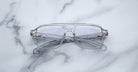 The Jacques Marie Mage Evans by Jacques Marie Mage are oversized acetate eyeglasses with clear frames and slightly tinted lenses, shown folded on a white marble surface.