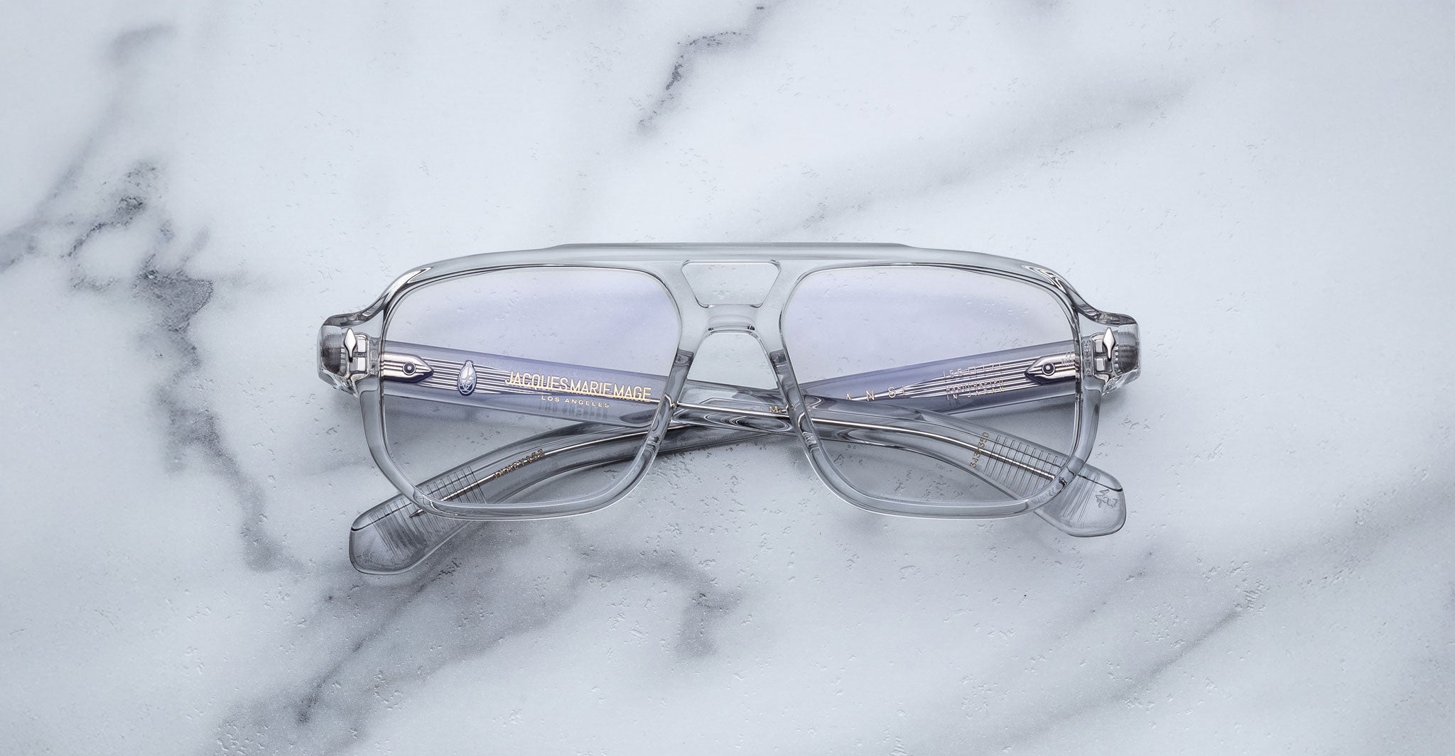 The Jacques Marie Mage Evans by Jacques Marie Mage are oversized acetate eyeglasses with clear frames and slightly tinted lenses, shown folded on a white marble surface.