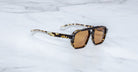 The Jacques Marie Mage Evans sunglasses feature an oversized tortoiseshell acetate frame, brown lenses, and clear arm tips. Displayed on white marble, they embody the refined style of Jacques Marie Mage.
