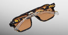 A pair of Jacques Marie Mage Evans sunglasses, featuring an oversized tortoiseshell acetate frame, brown lenses, and clear foldable arms, is folded and viewed from above against a light gray background.