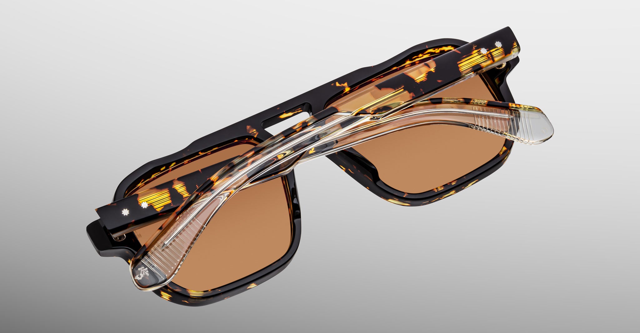 A pair of Jacques Marie Mage Evans sunglasses, featuring an oversized tortoiseshell acetate frame, brown lenses, and clear foldable arms, is folded and viewed from above against a light gray background.