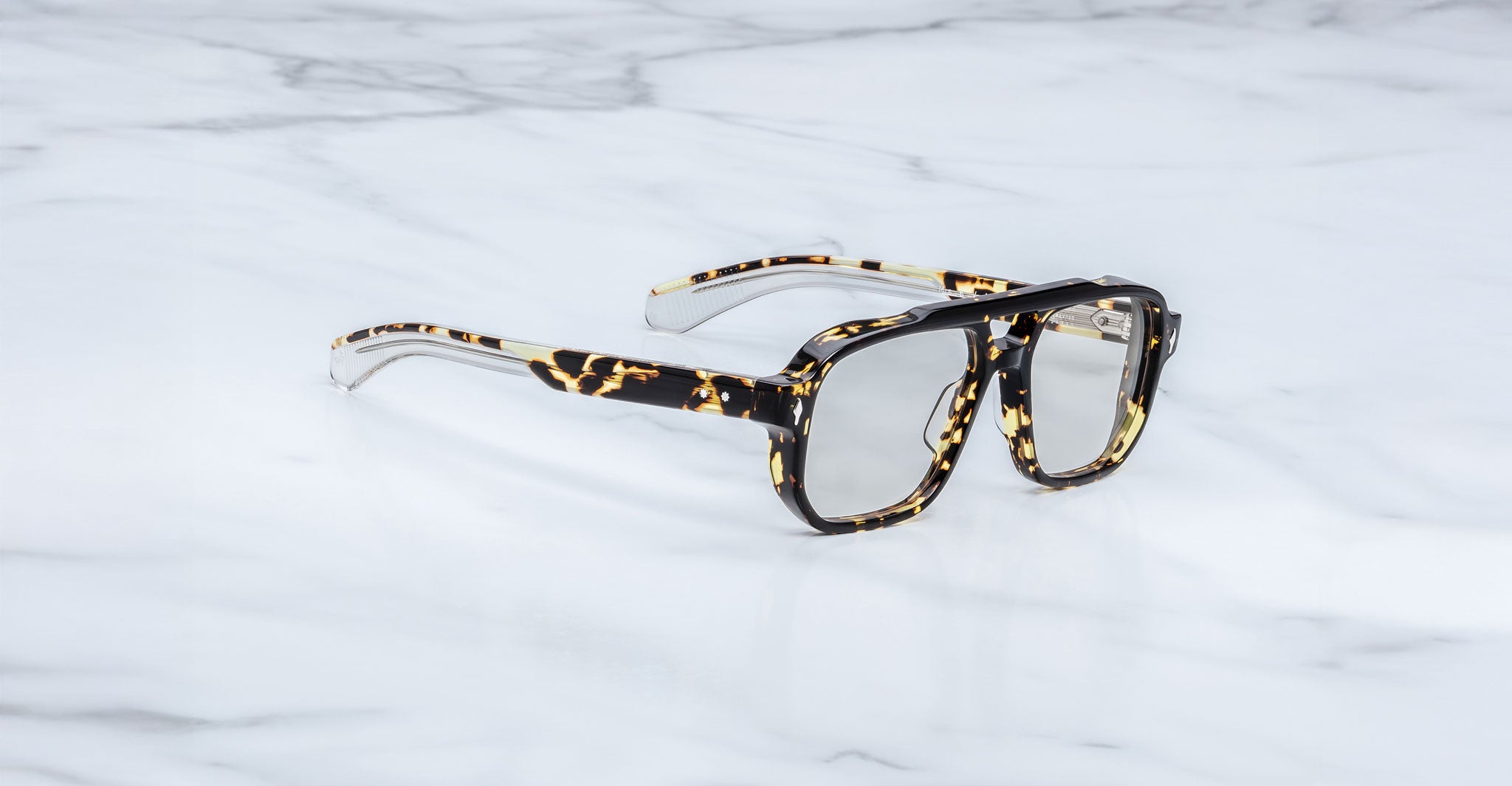 The Jacques Marie Mage Evans are handcrafted optical glasses with oversized tortoiseshell acetate frames and clear lenses, inspired by Jacques Marie Mage, shown resting on a white marble surface.