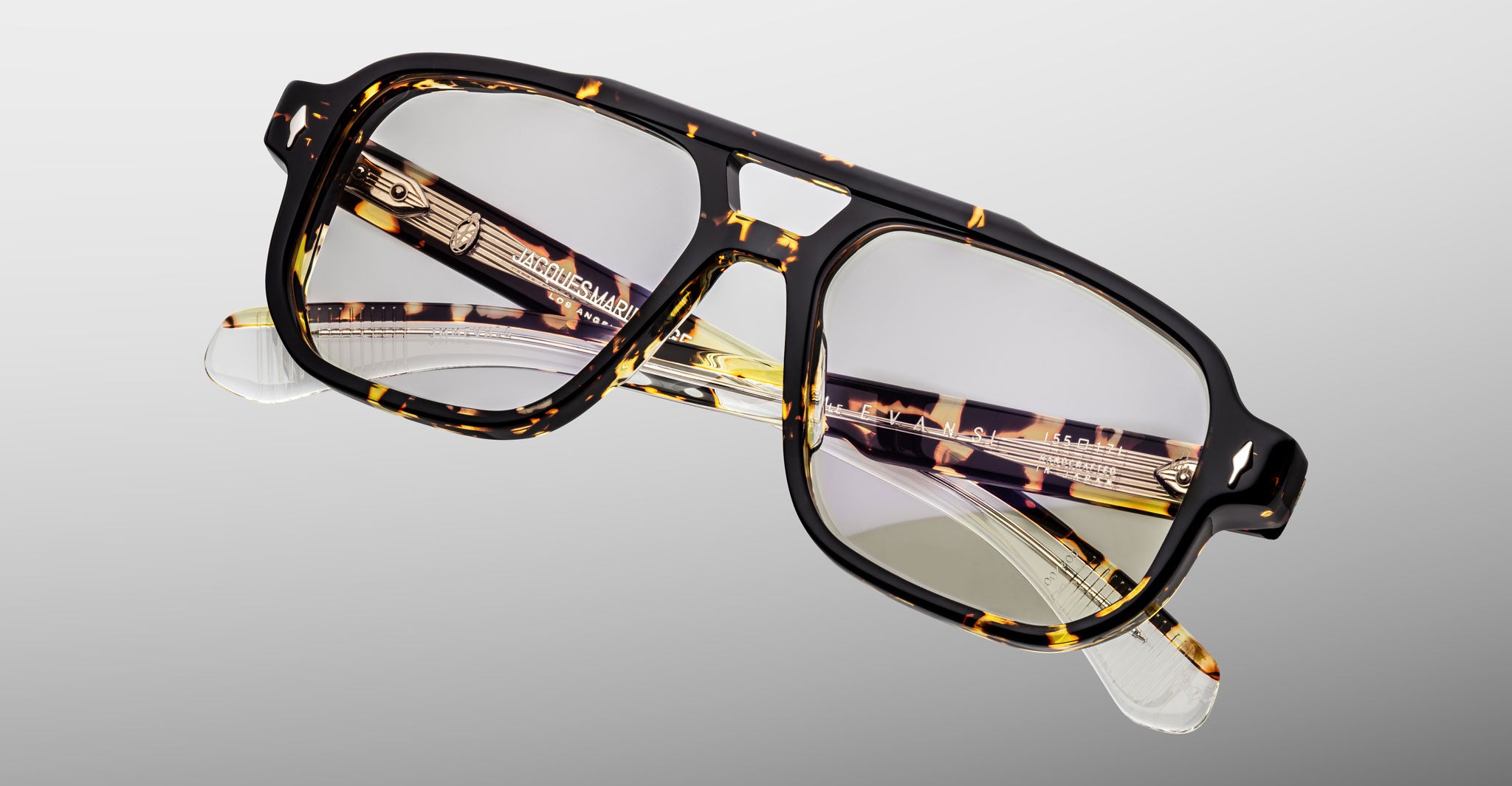 The Jacques Marie Mage Evans eyeglasses feature an oversized tortoiseshell acetate frame, clear lenses, and gold temple accents, presented against a light gray background.