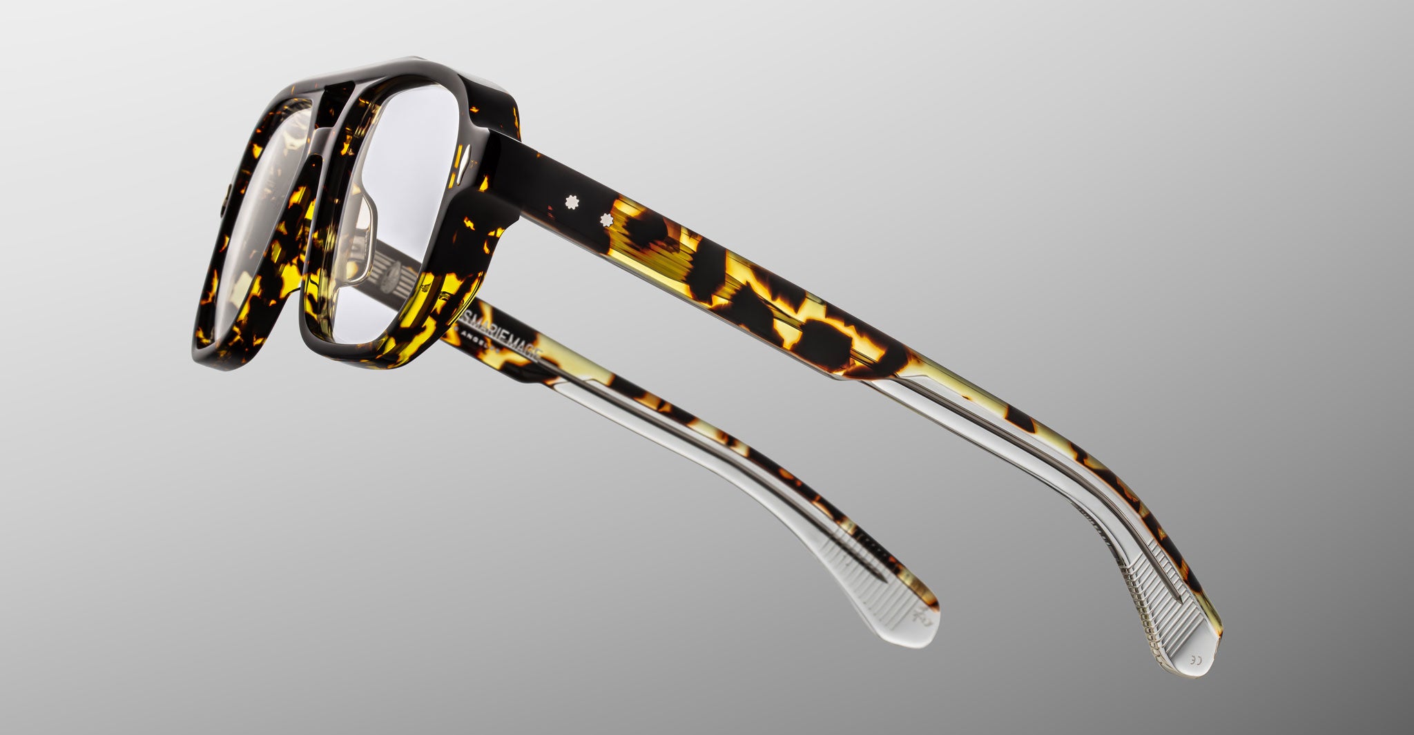 The Jacques Marie Mage Evans optical glasses showcase a handcrafted, oversized rectangular acetate frame in tortoiseshell with clear lower arms, set against a light gray background.