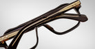 Close-up of folded Jacques Marie Mage Evans eyeglasses featuring black and gold oversized acetate frames, viewed from the side against a plain light background.