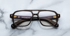 The Jacques Marie Mage Evans are thick-rimmed rectangular eyeglasses with black acetate frames and gold detailing by Jacques Marie Mage, displayed on a light marble surface.