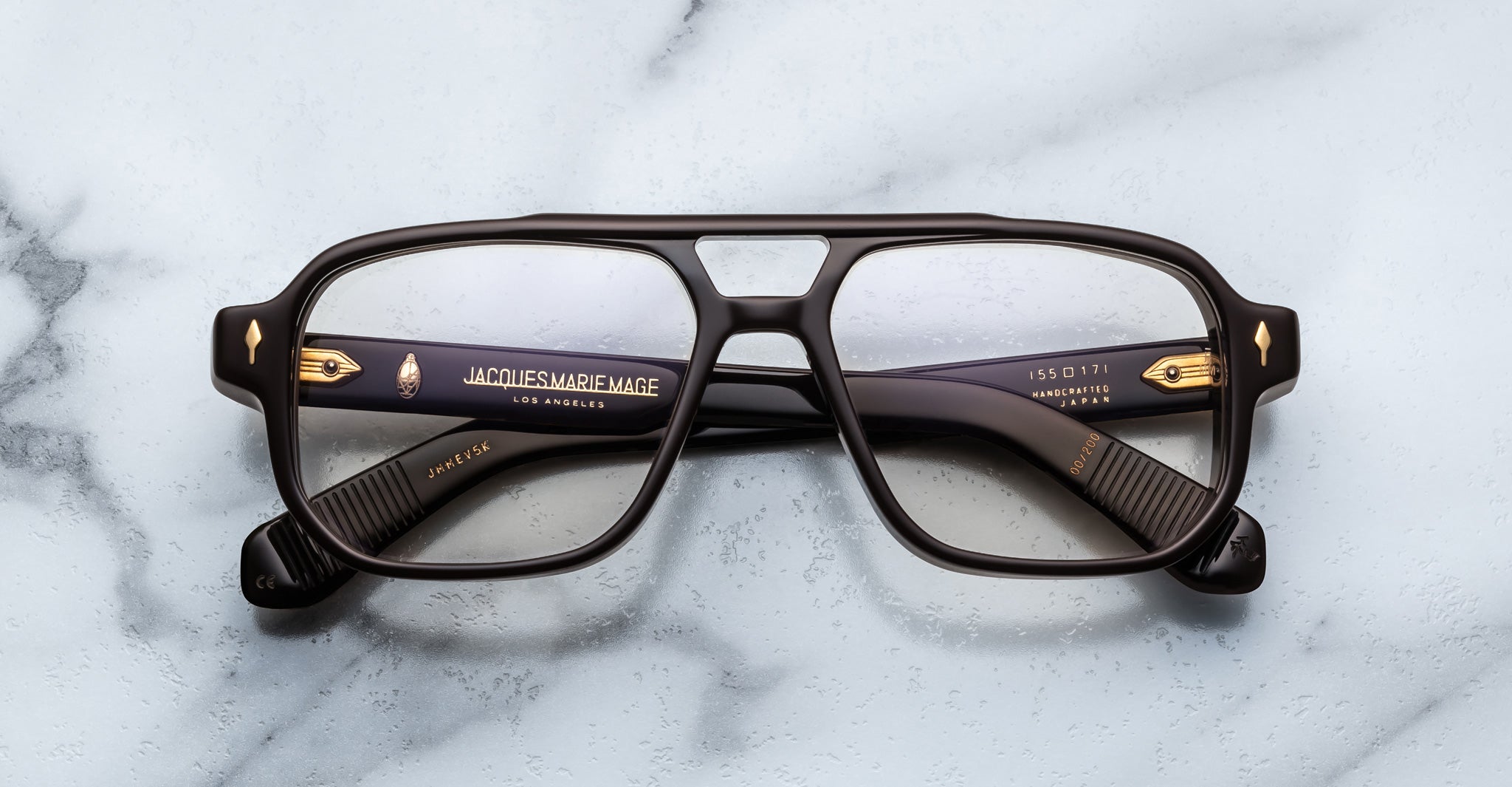 The Jacques Marie Mage Evans are thick-rimmed rectangular eyeglasses with black acetate frames and gold detailing by Jacques Marie Mage, displayed on a light marble surface.