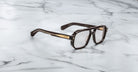 The Jacques Marie Mage Evans eyeglasses, featuring a bold rectangular acetate frame in black and gold, rest elegantly on a white marble surface.