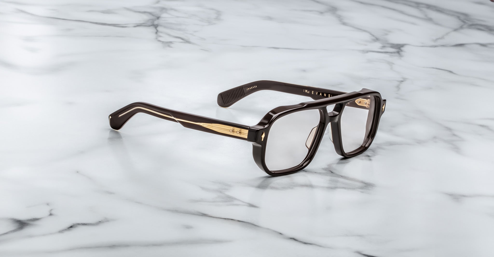 The Jacques Marie Mage Evans eyeglasses, featuring a bold rectangular acetate frame in black and gold, rest elegantly on a white marble surface.