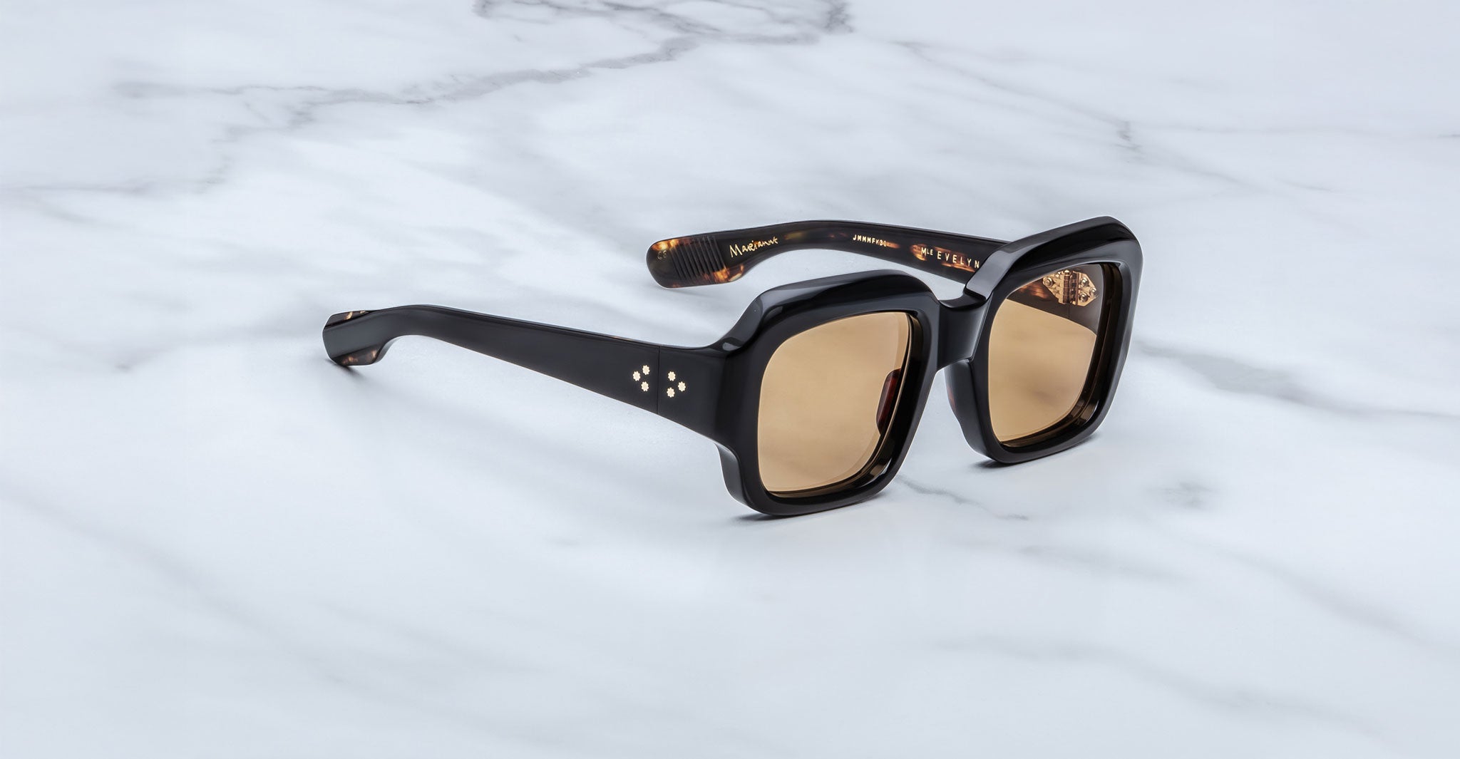 The Jacques Marie Mage Evelyn sunglasses, featuring black square frames and brown-tinted lenses, are displayed on a white marble surface. The arms include subtle gold accents and branding on the inside.