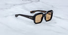 The Jacques Marie Mage Evelyn sunglasses, featuring black square frames and brown-tinted lenses, are displayed on a white marble surface. The arms include subtle gold accents and branding on the inside.