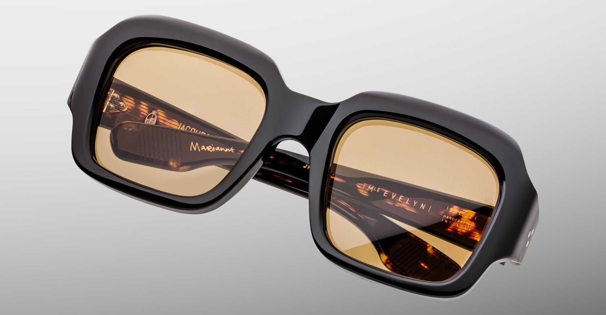 The Jacques Marie Mage Evelyn features oversized square black frames with amber-tinted lenses, displayed at an angle on a light gray background. The inside arms showcase gold branding and elegant tortoiseshell accents.