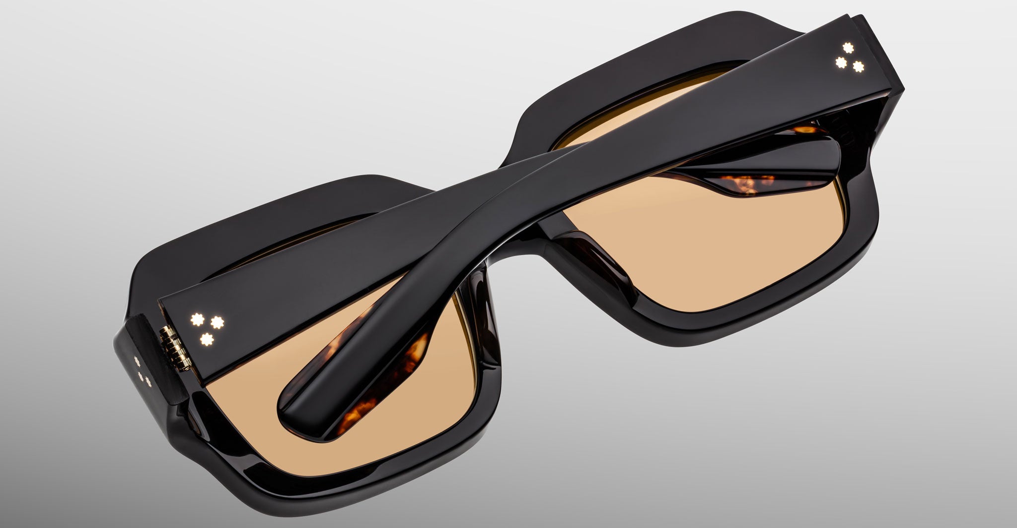 The Jacques Marie Mage Evelyn eyeglasses, featuring thick black geometric frames and amber-tinted lenses, are displayed upside down with crossed arms on a light gray background.