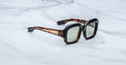 The Jacques Marie Mage Evelyn sunglasses feature a rectangular tortoiseshell design with thick frames and gold-accented arms, displayed on a white marble surface.