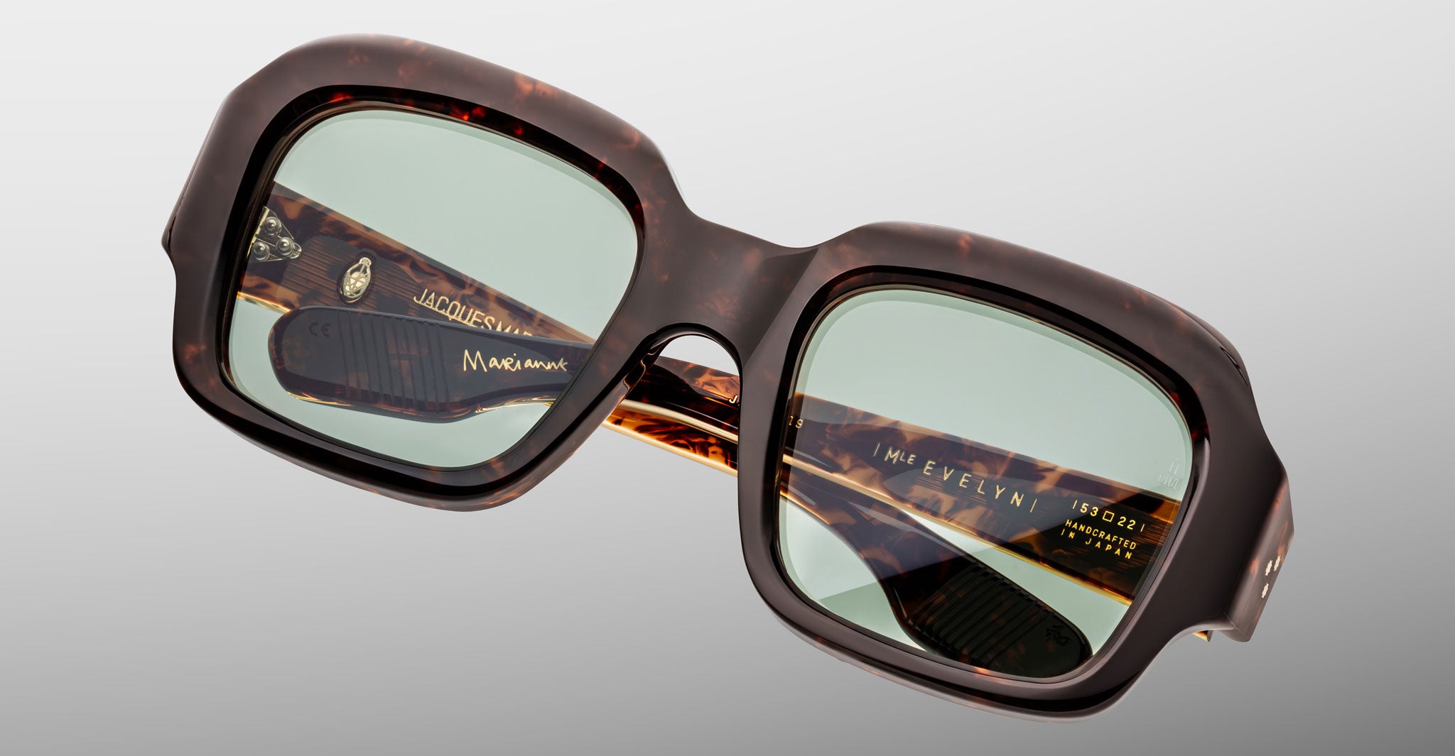 The Jacques Marie Mage Evelyn eyeglasses feature a square tortoiseshell frame with green-tinted lenses, shown against a light gray background. Gold branding and model details appear inside the temple arms.