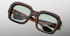 The Jacques Marie Mage Evelyn eyeglasses feature a square tortoiseshell frame with green-tinted lenses, shown against a light gray background. Gold branding and model details appear inside the temple arms.