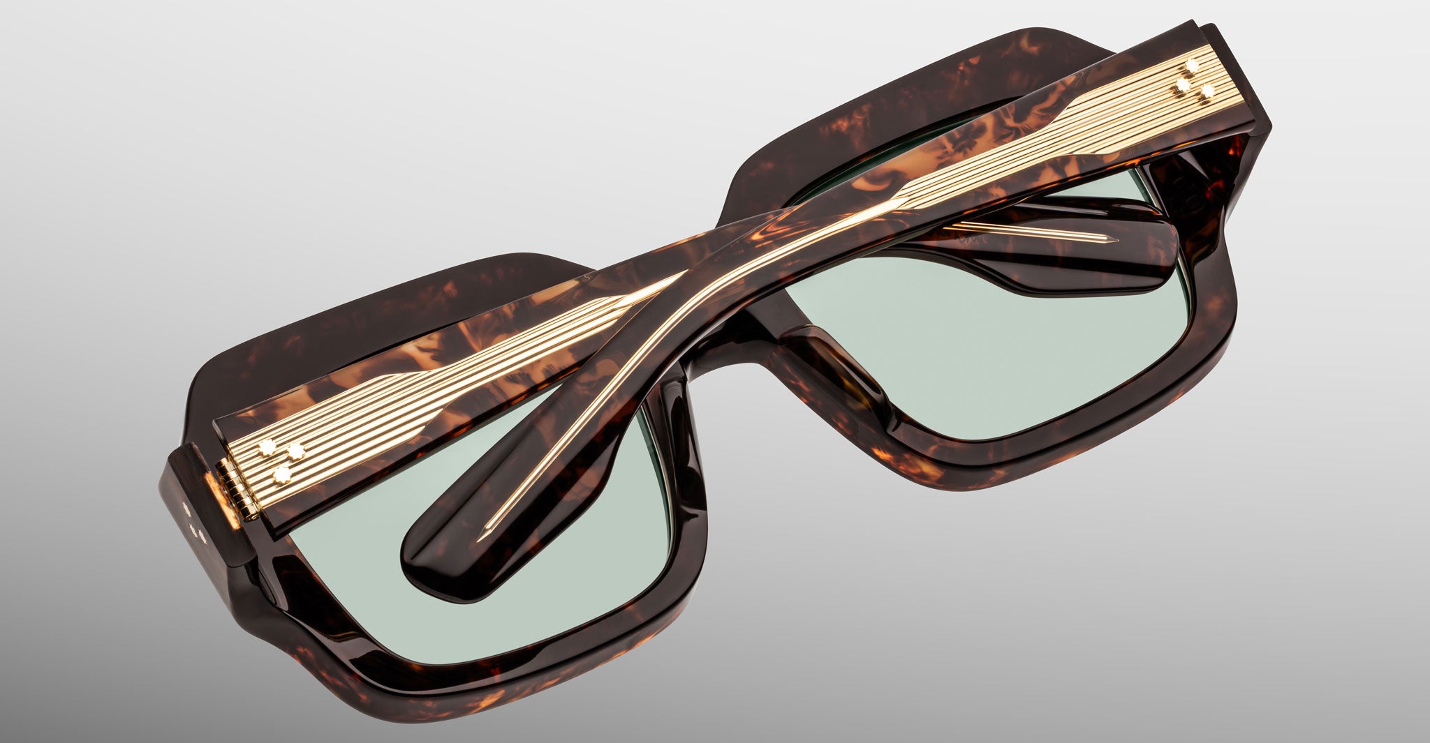 The Jacques Marie Mage Evelyn sunglasses are tortoiseshell and rectangular with green-tinted lenses, gold accents, and horizontal gold lines on the temples, shown folded closed.