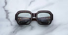 A pair of Jacques Marie Mage Evelyn eyeglasses featuring large, square, dark frames with thick rims rests face-up on a white marble surface, with the lens reflections revealing text inside the arms.