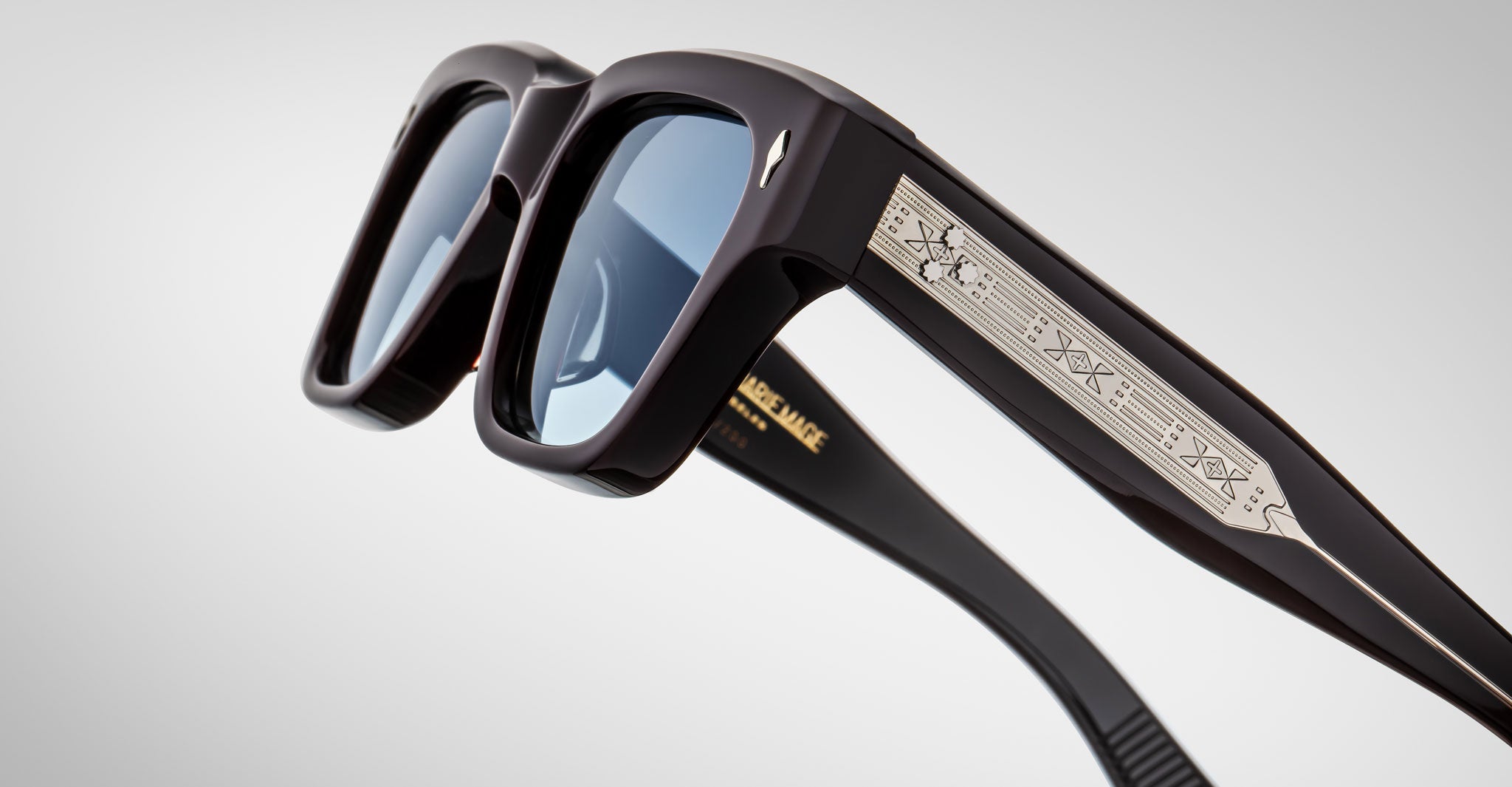 Close-up of Jacques Marie Mage Fellini sunglasses, black frame, blue lenses, metallic engraved arms, front and temple visible.