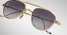 Close-up of Jacques Marie Mage Flynn gold-framed aviator sunglasses with dark tinted lenses, metal nose pads, and a brow bar set against a plain light grey background.