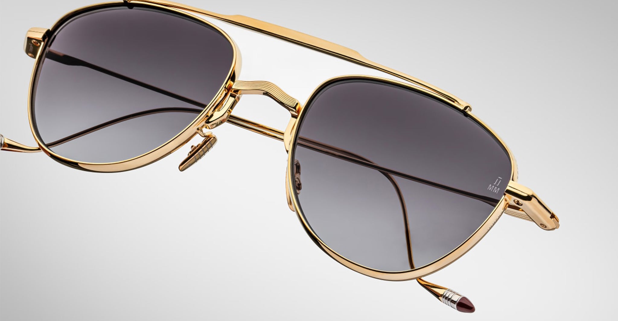 Close-up of Jacques Marie Mage Flynn gold-framed aviator sunglasses with dark tinted lenses, metal nose pads, and a brow bar set against a plain light grey background.