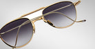 A close-up of Jacques Marie Mage Flynn gold-framed aviator sunglasses with dark gradient lenses, tilted to showcase the frame and temples, set against a plain light gray background.