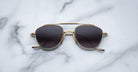 A pair of Jacques Marie Mage Flynn gold-framed sunglasses with dark lenses rests on a white marble surface accented by gray veining.