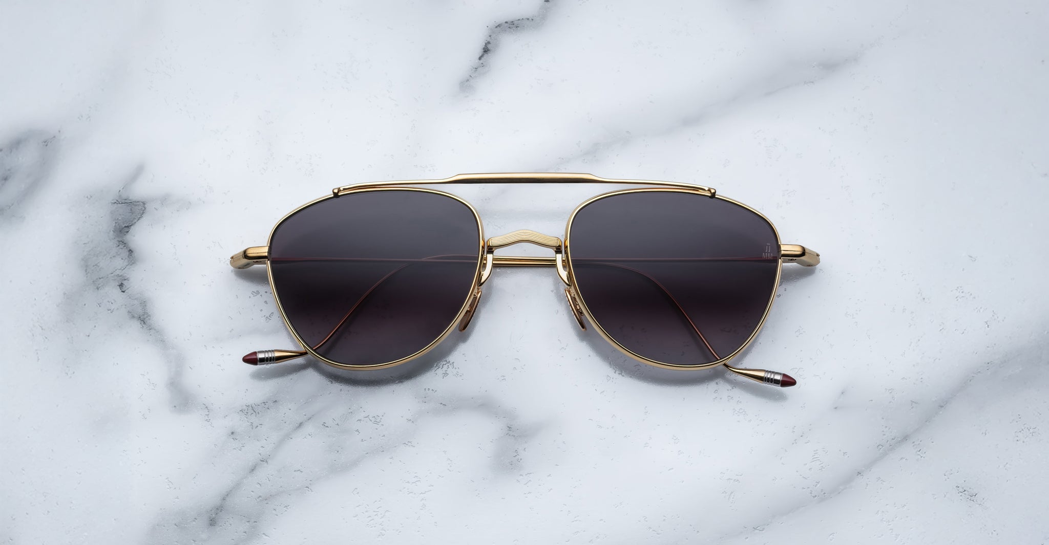 A pair of Jacques Marie Mage Flynn gold-framed sunglasses with dark lenses rests on a white marble surface accented by gray veining.