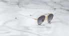 The Jacques Marie Mage Flynn sunglasses, featuring gold frames and dark reflective lenses, are displayed at an angle on a polished white marble surface with gray veining, showcasing their sleek, thin metal arms.
