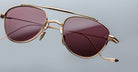 A close-up of Jacques Marie Mage Flynn gold-framed aviator sunglasses with reddish-brown tinted lenses, set against a plain light gray background. The frame details and reflections are clearly visible.