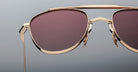 Close-up of Jacques Marie Mage Flynn sunglasses with gold frames, pink-tinted lenses, and a double bridge design, set against a plain gray background.